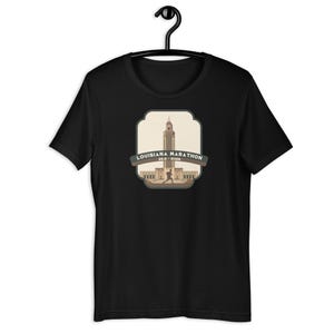 May include: Black t-shirt with a graphic of the Louisiana State Capitol building and the text "Louisiana Marathon 2023 - 2026".