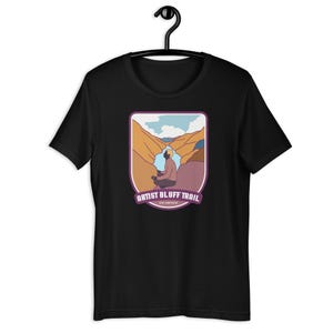 Puede incluir: Camiseta negra con un gráfico de una persona meditando en un cañón. El diseño incluye el texto "ARTIST BLUFF TRAIL" en una pancarta morada. La camiseta está colgada en una percha negra.