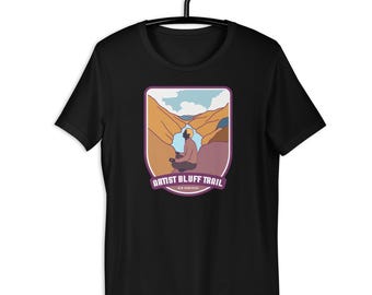 May include: Black t-shirt featuring a graphic of a person meditating in a canyon landscape. The design includes the text "ARTIST BLUFF TRAIL" in a purple banner. The shirt is hanging on a black hanger.