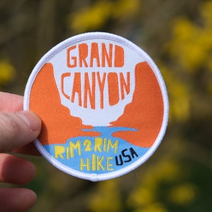May include: A round patch with the words "GRAND CANYON" in orange block letters. Below, the text "RIM 2 RIM HIKE USA" is in blue and yellow. The patch features an orange canyon design and a white border.