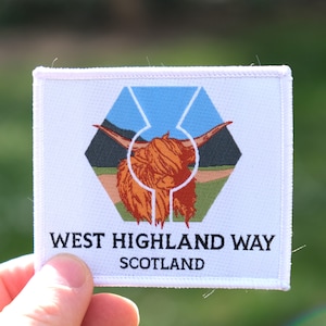 Könnte beinhalten: Weißer Patch mit einem sechseckigen Logo, das eine rote Highland-Kuh mit blauem Himmel und grünen Hügeln im Hintergrund zeigt. Der Text "WEST HIGHLAND WAY SCOTLAND" ist unter dem Logo gedruckt.