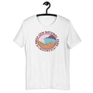 Puede incluir: Camiseta blanca con un gráfico circular marrón con el texto "Kawah Ijen National Park" y una ilustración estilizada de una cordillera con un lago azul.
