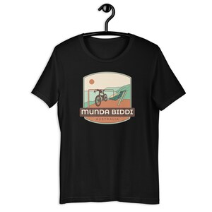 May include: Black t-shirt featuring a graphic of a bicycle on a trail with the text "Munda Biddi Australia". The design includes a sun, a bicycle, and a trail scene in shades of brown, green, and orange.