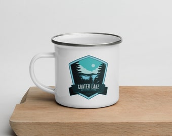 May include: White enamel camping mug with a blue and green graphic of Crater Lake National Park, Oregon. The mug has a silver rim and handle.