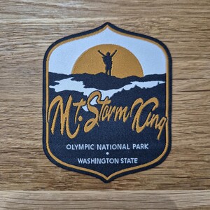 Puede incluir: Un parche bordado en negro y dorado con una silueta de una persona con los brazos levantados frente a un sol amarillo. El texto "Mt. Storm King" está en escritura dorada. Los textos "Olympic National Park" y "Washington State" están en blanco.