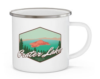 May include: White enamel mug with a graphic of Crater Lake National Park in Oregon. The graphic features a stylized image of the lake with a mountain in the background and green trees.