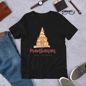 Op de afbeelding: Zwart T-shirt met een afbeelding van de Prambanan-tempel in Java, Indonesië. De tempel is getekend in een cartoon-stijl met oranje en gele kleuren. De tekst "Prambanan" is geschreven in roze cursieve letters onder de tempel.