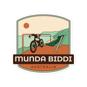 May include: A sticker featuring a scenic illustration of a bicycle parked near a bench with a coastal backdrop. The text "Munda Biddi Australia" is displayed in a brown rectangular banner at the bottom, with a light green border.