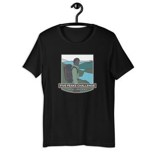 Pode incluir: Camiseta preta com um gráfico de uma pessoa fazendo caminhadas com uma mochila na frente de uma cordilheira. O gráfico é cercado por uma borda azul e branca com o texto "Five Peaks Challenge UK & Ireland".