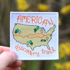 Op de afbeelding: Witte patch met oranje tekst "AMERICAN discovery trail" en een kaart van de Verenigde Staten met een rode lijn eroverheen en groene bomen.