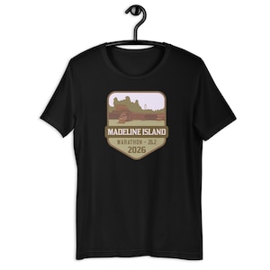 May include: Black t-shirt with a brown and green badge featuring a landscape scene and the text "Madeline Island Marathon - 26.2 2026".