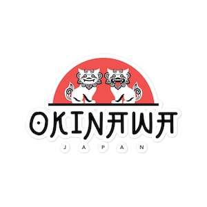 May include: A white sticker with a red sun and two black and white lion-like figures. The text "OKINAWA JAPAN" is written in black below the figures.
