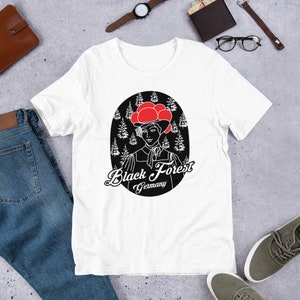 May include: White t-shirt featuring a black and white illustration of a woman in traditional Black Forest attire, with a red hat. The text "Black Forest Germany" is below the illustration. The shirt is made of a soft material.