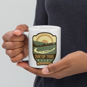 May include: White ceramic mug with a graphic of a mountain trail with a sun and trees. The text "SunTop Trail #1183 Washington" is printed on the mug.