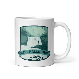 May include: White ceramic coffee mug with a green and blue illustration of a waterfall and a person standing on a rock. The text "Ousel Falls Trail MT" is printed below the illustration.