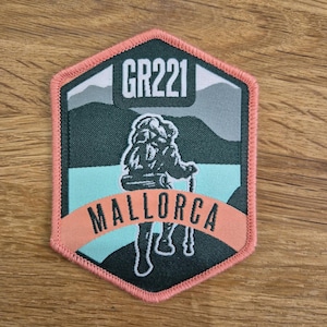 May include: A hexagonal shaped patch with a hiker silhouette in the centre. The patch is green, blue, and pink with the text "GR221" and "MALLORCA" on it.