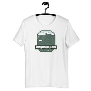 May include: White t-shirt featuring a green graphic of Bromo Tengger Semeru National Park in Indonesia. The graphic depicts a mountain range with a blue sky and clouds.