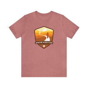 Puede incluir: Una camiseta rosa polvorienta con un diseño de paisaje desértico. El gráfico incluye un ciclista en un camino, un sol y el texto "Apache Wash Loop Cave Creek AZ" dentro de un emblema en forma de escudo. La camiseta es de material suave.