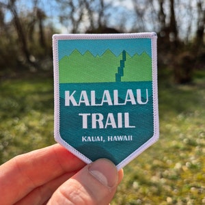 Pode incluir: Um remendo bordado azul-turquesa e branco com o texto "Kalalau Trail" e "Kauai, Hawaii". O remendo apresenta uma cordilheira verde com uma cachoeira.