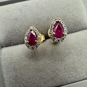 May include: A pair of gold earrings featuring pear-shaped ruby gemstones set within a halo of smaller diamonds.