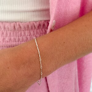 May include: A delicate silver chain bracelet with a simple design. The bracelet is made of small, linked pieces of metal that are slightly elongated. The bracelet is shown on a person's wrist.