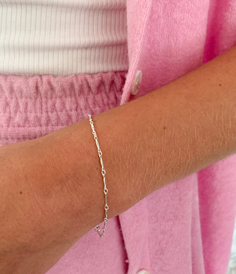 May include: A delicate silver chain bracelet with a simple design. The bracelet is made of small, linked pieces of metal that are slightly elongated. The bracelet is shown on a person's wrist.