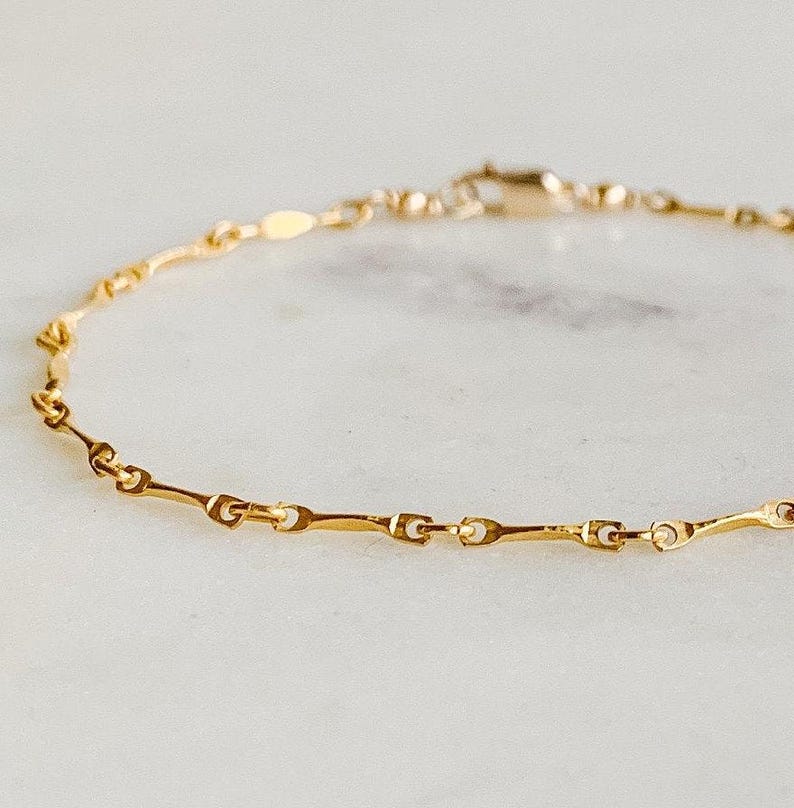May include: A delicate gold chain bracelet with a simple, elegant design. The chain is made of small, linked pieces that create a subtle wave pattern.