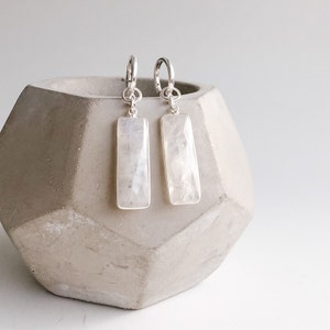 May include: A pair of drop earrings featuring rectangular, faceted moonstone gems set in silver frames. Each stone is suspended from a small silver hoop by a circular jump ring, showcasing a translucent, milky white appearance with subtle iridescence.
