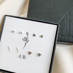 May include: A white box with a black border containing six pairs of silver earrings. The earrings are in the shapes of circles, hearts, and triangles. The box also has the letters "A/R" printed on it.