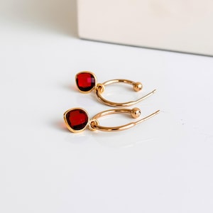 May include: A pair of gold-coloured hoop earrings, each with a square, deep red gemstone. The earrings are presented on a white surface, with a hint of a white background.