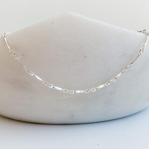 May include: A delicate silver chain necklace with a flat, oval-shaped link design.