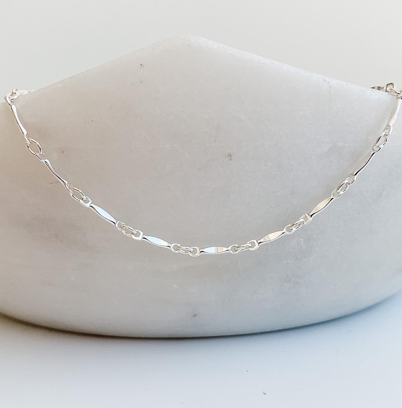 May include: A delicate silver chain necklace with a flat, oval-shaped link design.