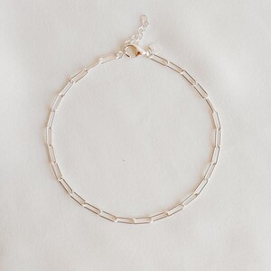 May include: A delicate silver paperclip chain bracelet. The bracelet features a series of elongated, rectangular links connected to form a complete circle. The clasp is a lobster claw style. The jewelry is displayed on a white background.