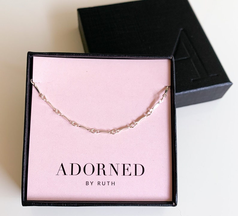 May include: A silver chain necklace in a pink box with the text "ADORNED BY RUTH".