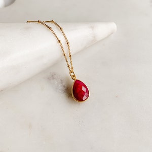 May include: A gold chain necklace featuring a faceted, teardrop-shaped ruby pendant set in a gold bezel. The chain is composed of small, evenly spaced gold beads, and the deep red gemstone has a textured, multi-faceted surface that reflects light.