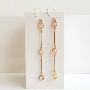 May include: A pair of gold-tone drop earrings. Each earring has a fine gold wire hook, linked to three clear, round gemstones. The gemstones are set along a slender gold bar, creating an elegant, linear design. The earrings are a stylish accessory.