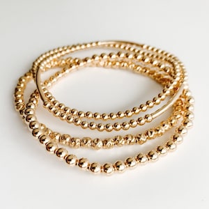 May include: Four gold beaded bracelets stacked on top of each other. The bracelets are made of small, round beads.