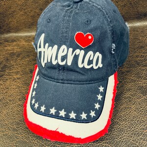 May include: A black baseball cap with a white "America" logo and a red heart. The brim of the cap has a white band with white stars and a red frayed edge.