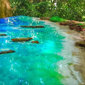Może przedstawiać: Zbliżenie na dzieło sztuki z żywicy przedstawiające scenę plażową. Dzieło przedstawia turkusowy ocean z białymi, pienistymi falami, piaszczystą plażę i brązowe dryfujące drewno. Dzieło jest umieszczone na tle zielonej roślinności.