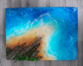 May include: A resin art piece depicting a beach scene with blue water, white foam, and a sandy shore. The bottom of the piece features a piece of driftwood with green moss.