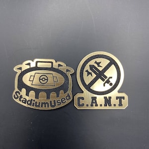 May include: Two gold-colored metal pins. The first pin features a black and white illustration of a stadium with the text "Stadium Used". The second pin features a black and white illustration of a sword with a circle around it and the text "C.A.N.T."