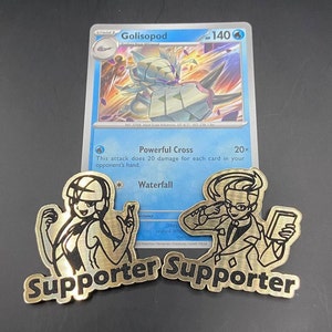 May include: Two gold metal Pokemon supporter cards. The cards feature black and white illustrations of characters from the Pokemon Trading Card Game. The text "Supporter" is printed in black on each card.