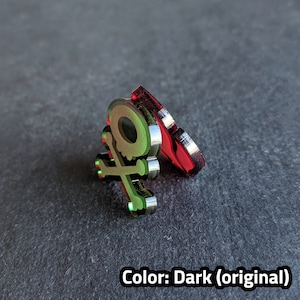 Puede incluir: Un dise&ntilde;o de calavera y huesos cruzados verde y plateado sobre una base de acr&iacute;lico rojo. El dise&ntilde;o est&aacute; en relieve y tiene un acabado met&aacute;lico. Color: Oscuro (original)