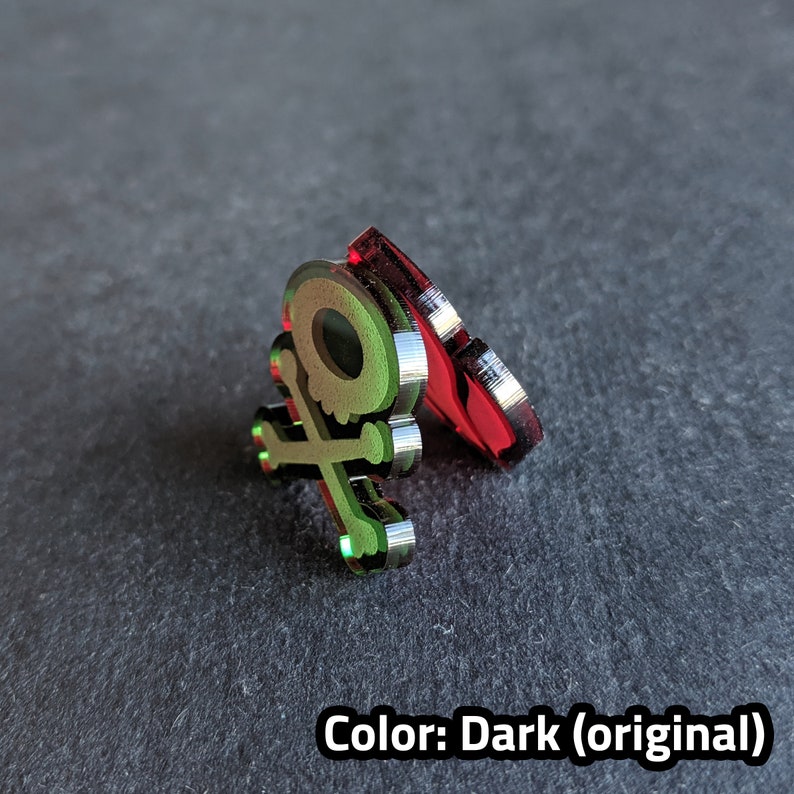 Puede incluir: Un dise&ntilde;o de calavera y huesos cruzados verde y plateado sobre una base de acr&iacute;lico rojo. El dise&ntilde;o est&aacute; en relieve y tiene un acabado met&aacute;lico. Color: Oscuro (original)