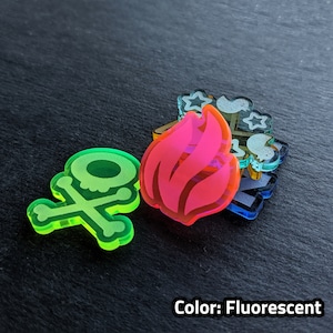 Puede incluir: Un conjunto de cuatro fichas acr&iacute;licas fluorescentes que presentan una calavera y huesos cruzados, una llama y otros dos dise&ntilde;os abstractos. Las fichas est&aacute;n superpuestas.