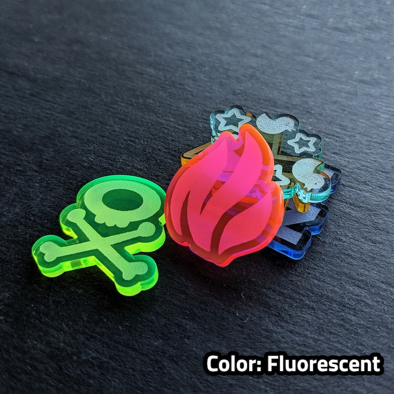 Puede incluir: Un conjunto de cuatro fichas acr&iacute;licas fluorescentes que presentan una calavera y huesos cruzados, una llama y otros dos dise&ntilde;os abstractos. Las fichas est&aacute;n superpuestas.