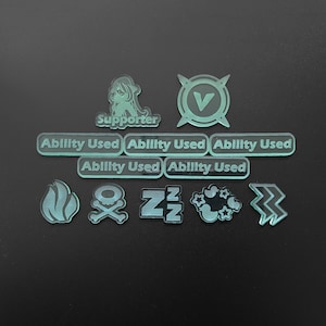 May include: A collection of translucent light green acrylic tokens featuring various icons and text. The set includes five rectangular tokens labeled Ability Used, a Supporter token, a V-logo token, and icons for fire, skull and crossbones, ZZZ, ducks, and lightning.