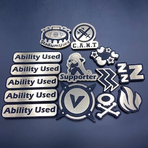 May include: A collection of black and gold Pokemon trading card game tokens. The tokens include a stadium, a supporter, a 'C.A.N.T' token, a 'Z' token, a 'V' token, a skull and crossbones token, a flame token, and five 'Ability Used' tokens.