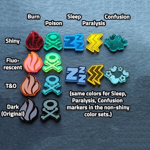 Puede incluir: Cuatro filas de marcadores de juego de pl&aacute;stico de colores. La fila superior muestra una llama roja, una calavera y huesos cruzados verdes, un s&iacute;mbolo 'ZZ' azul, un rayo amarillo y una nube turquesa con tres p&aacute;jaros. La segunda fila muestra una llama rosa, una calavera y huesos cruzados verdes, un s&iacute;mbolo 'ZZ' azul claro, un rayo amarillo y una nube turquesa con tres p&aacute;jaros y estrellas. La tercera fila muestra una llama naranja, una calavera y huesos cruzados naranja, un s&iacute;mbolo 'ZZ' azul claro, un rayo amarillo y una nube turquesa con tres p&aacute;jaros y estrellas. La fila inferior muestra una llama gris, una calavera y huesos cruzados grises, un s&iacute;mbolo 'ZZ' azul claro, un rayo amarillo y una nube turquesa con tres p&aacute;jaros y estrellas. El texto 'Shiny', 'Fluo-rescent', 'T&O' y 'Dark (Original)' est&aacute; escrito a la izquierda de cada fila. El texto 'Burn', 'Poison', 'Sleep', 'Paralysis' y 'Confusion' est&aacute; escrito encima de la fila superior. El texto '(mismos colores para Sleep, Paralysis, Confusion markers en los conjuntos de colores no brillantes.)' est&aacute; escrito debajo de la tercera fila.
