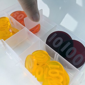 Puede incluir: Un contenedor de almacenamiento de plástico transparente con una variedad de fichas de plástico de colores. Las fichas están numeradas 100, 101 y 102. El contenedor tiene una tapa transparente y está abierto por un lado.